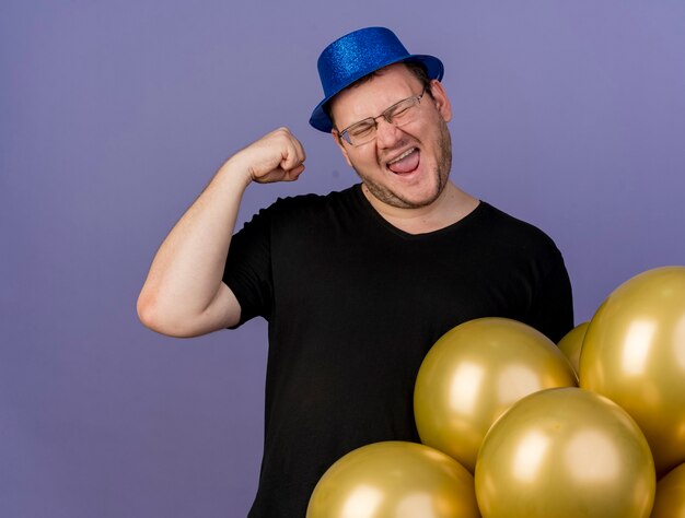 Felice uomo slavo adulto in occhiali ottici che indossa un cappello da festa blu alza il pugno in piedi con palloncini di elio