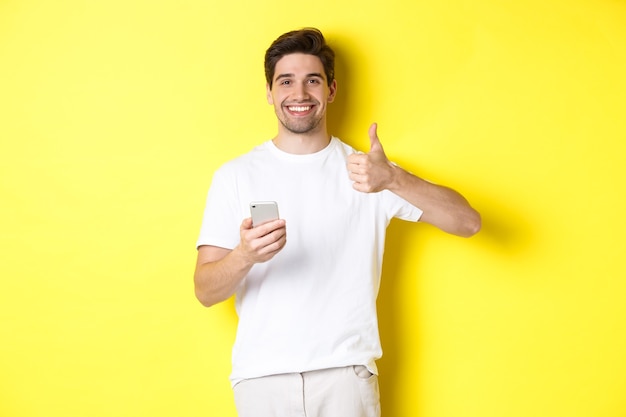 Felice uomo soddisfatto che tiene in mano lo smartphone, mostra il pollice in su in segno di approvazione, consiglia qualcosa online, in piedi su sfondo giallo