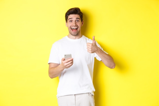 Felice uomo sorridente che tiene in mano lo smartphone, mostra il pollice in su in segno di approvazione, consiglia qualcosa online, in piedi su sfondo giallo.