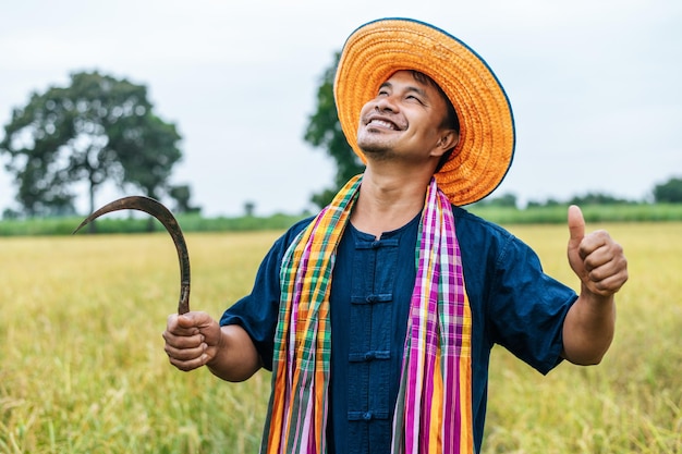 Felicemente giovane agricoltore che indossa cappello di paglia e perizoma in piedi con un colpo in su e tiene la falce nel campo di riso