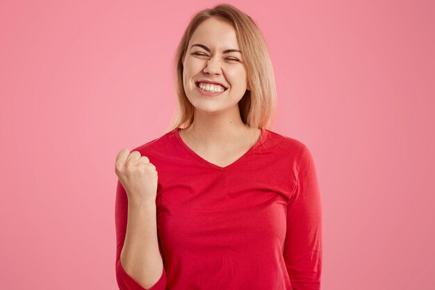 Felicissima donna europea con i capelli corti, stringe il pugno, vestita in abito casual rosso, viso strabico, vestita di maglione rosso