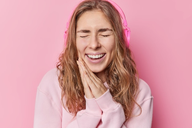 Felicissima ragazza dai capelli lunghi tiene le mani premute insieme ride con gioia chiude gli occhi ascolta musica tramite cuffie stereo wireless indossa una felpa casual isolata su sfondo rosa
