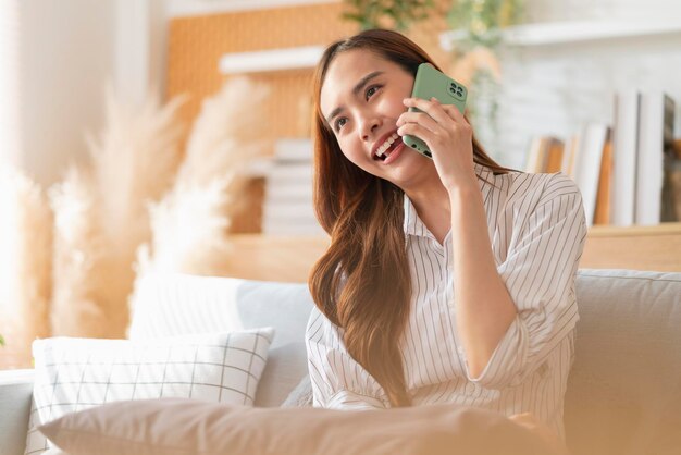 Felicità allegra sorridente giovane donna asiatica adulta donna indossa panno casual conversazione a mano sullo smartphone sul divano a casa quarantena remota periodo di blocco sfondo interno domestico