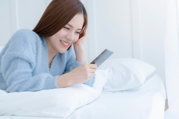 Felicità bella donna asiatica sorriso risveglio mattutino sul letto bianco cuscino morbido con sfondo camera da letto smartphone tenere in mano