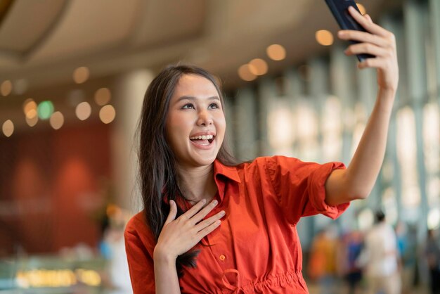 Felicità donna asiatica sorriso mano onda dare un bacio alla famiglia tramite videochiamata dispositivo smartphone saluto sociale distanza con sfocatura centro commerciale sfondo nuovo stile di vita normale
