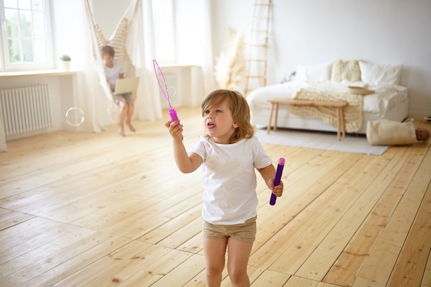 Felicità e concetto di infanzia spensierata. Ritratto dell'interno di adorabile bambino femmina felice in t-shirt e pantaloncini in piedi nel mezzo del soggiorno moderno, divertendosi, soffiando bolle di sapone,