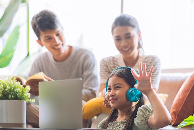 Felicità famiglia asiatica figlia studio online da casa con il laptop mentre padre madre è seduto insieme guarda con sorriso allegro bambino asiatico femmina indossare cuffie rispondere alla domanda dell'insegnante