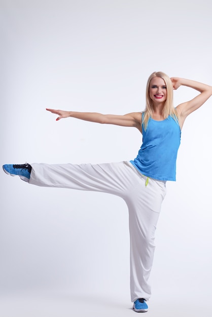 femmina bionda in attrezzatura hip-hop di dancing che equilibra su una gamba che sorride sul bianco