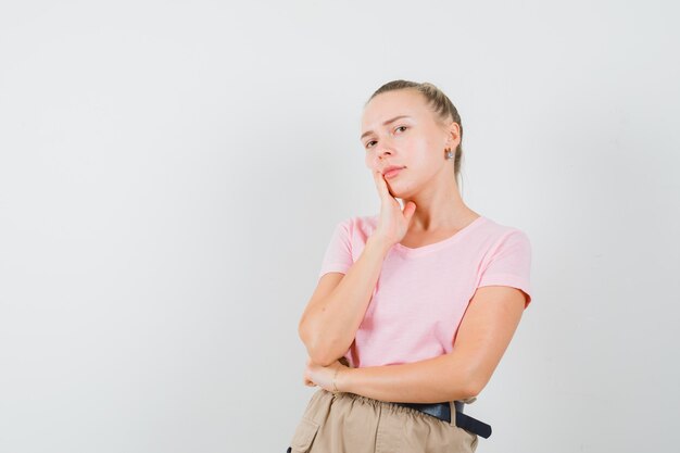 Femmina bionda in piedi nel pensiero posa in t-shirt, pantaloni e sembra sensibile, vista frontale.