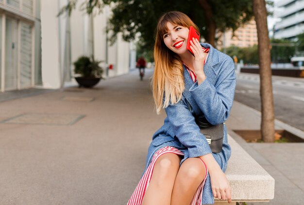 Femmina bionda sorridente riuscita che parla dal telefono cellulare sul fondo urbano dopo il lavoro. Seduto su una panchina.