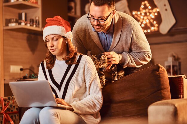 Femmina che celebra la vigilia di Natale a casa con il gatto. Coppia sposata utilizzando un laptop.