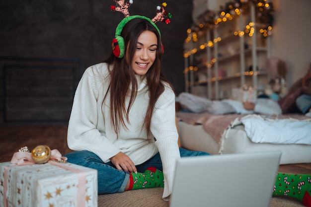 Femmina con corna sorridente mentre parla con un amico in linea sul computer portatile durante la celebrazione del Natale a casa