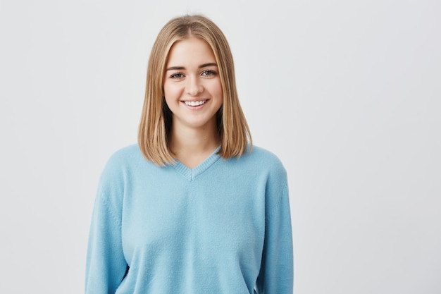 Femmina europea affascinante, felice, sorridente con aspetto attraente e capelli chiari che indossa un maglione blu che mostra i suoi denti perfetti con buon umore, godendo di posa.