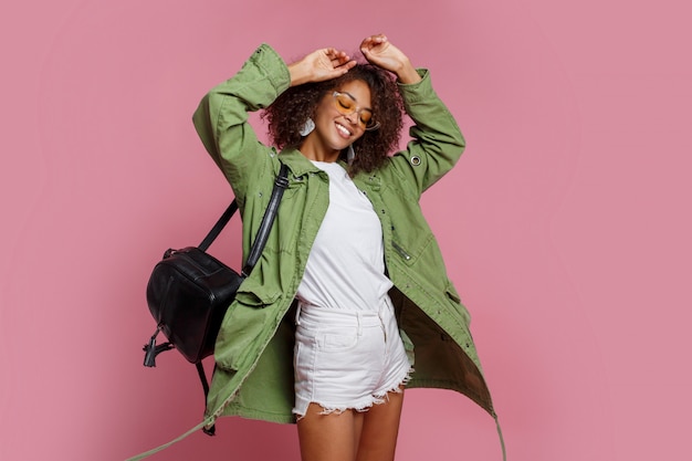 Femmina nera di Jocund in rivestimento alla moda della molla che posa nello studio su fondo rosa.