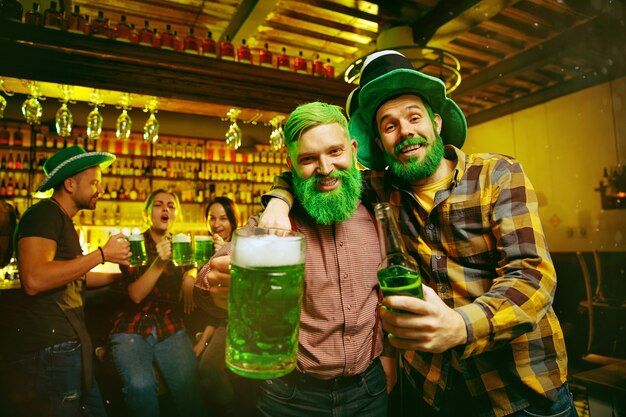 Festa di San Patrizio. Amici felici festeggia e beve birra verde. Giovani uomini e donne che indossano cappelli verdi. Pub interno.