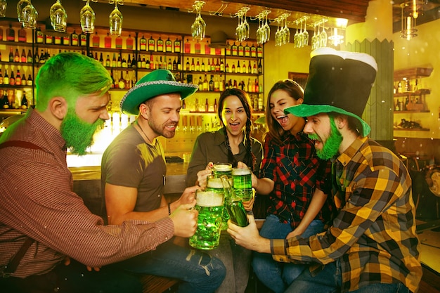 Festa di San Patrizio. Gli amici felici stanno festeggiando e bevendo birra verde. Giovani uomini e donne che indossano cappelli verdi. Pub interno.