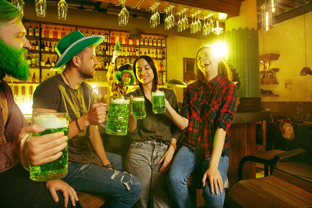 Festa di San Patrizio. Gli amici felici stanno festeggiando e bevendo birra verde. Giovani uomini e donne che indossano cappelli verdi. Pub interno.