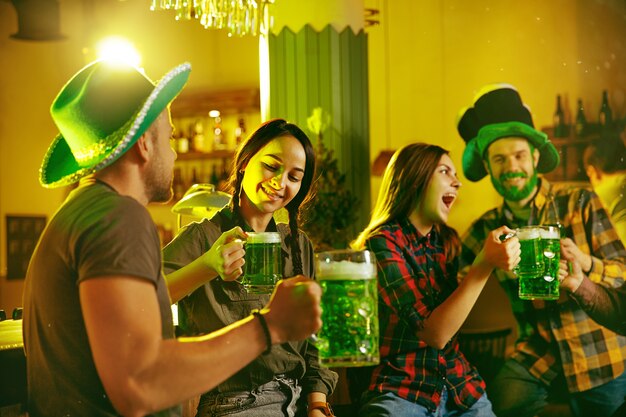 Festa di San Patrizio. Gli amici felici stanno festeggiando e bevendo birra verde. Giovani uomini e donne che indossano cappelli verdi. Pub interno.