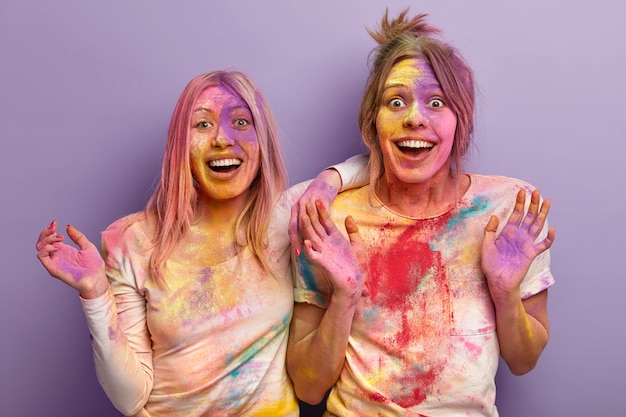 Festival di Holi, divertimento e concetto di persone. Due donne felicissime giocano con i colori, mostrano le palme, imbrattate di polvere colorata, isolate su un muro viola. Esplosione multicolore