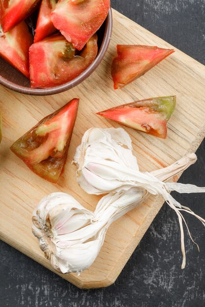 Fette del pomodoro con i bulbi dell'aglio in un piatto dell'argilla sulla parete del tagliere e di lerciume, disposizione piana.