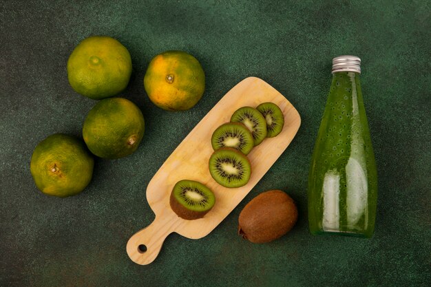 Fette di kiwi vista dall'alto su un tagliere con mandarini e una bottiglia di succo su una parete verde