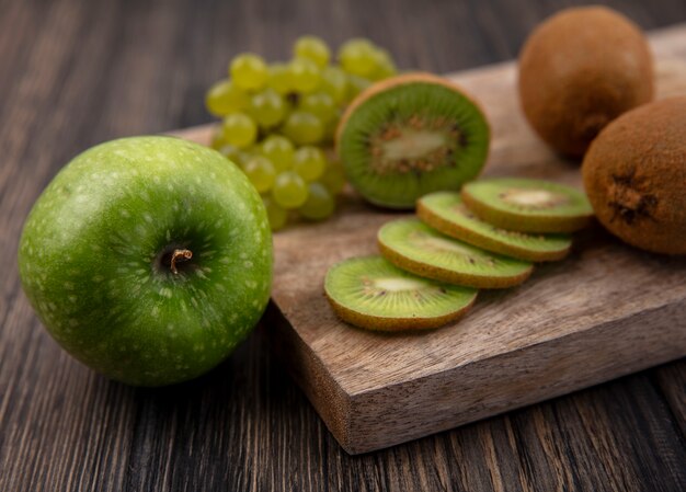 Fette di kiwi vista laterale con uva verde su un supporto con una mela verde su uno sfondo di legno