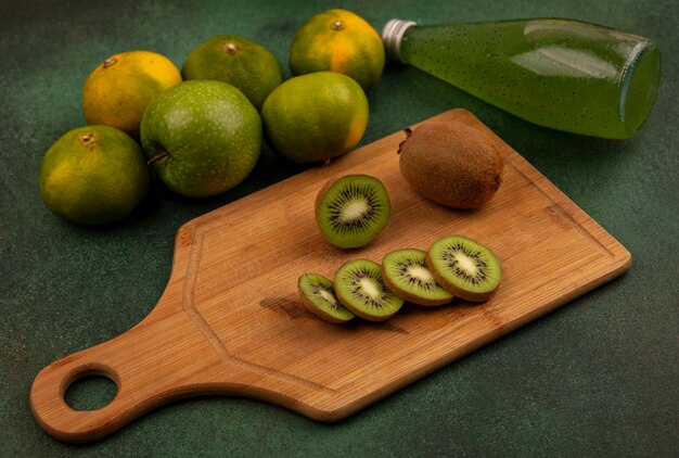 Fette di kiwi vista laterale su un tagliere con mandarini e una bottiglia di succo su una parete verde