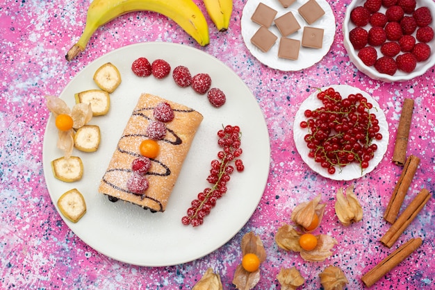 Fette di torta rotolo vista dall'alto con diversi frutti all'interno del piatto bianco sul colore dolce biscotto sfondo colorato