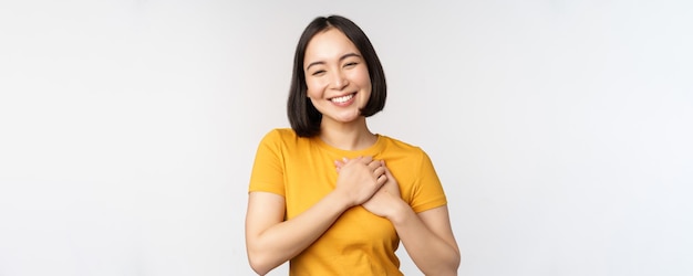 Fidanzata asiatica romantica che si tiene per mano sul cuore che sorride con cura e tenerezza che sta in maglietta gialla sopra fondo bianco
