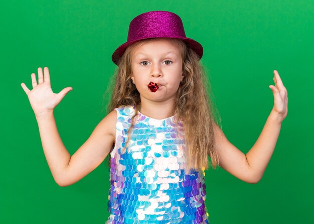 fiduciosa bambina bionda con cappello da festa viola in piedi con le mani alzate che soffia fischio di festa isolato sulla parete verde con spazio di copia