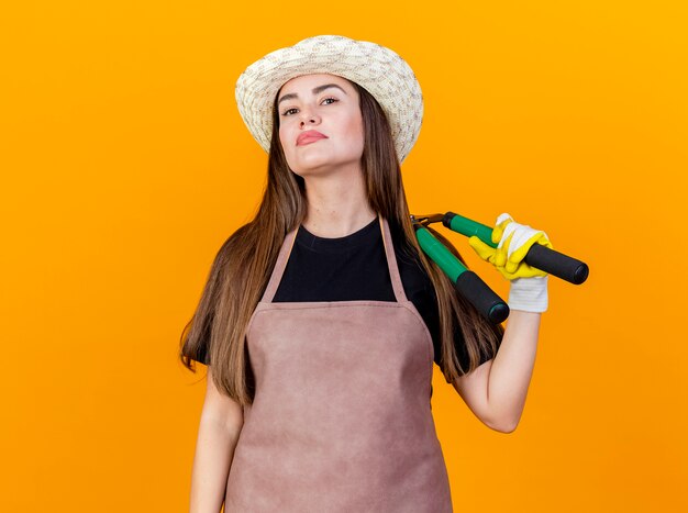 Fiduciosa bella ragazza giardiniere che indossa uniforme e cappello da giardinaggio con guanti che tengono i clippers sulla spalla isolati su sfondo arancione