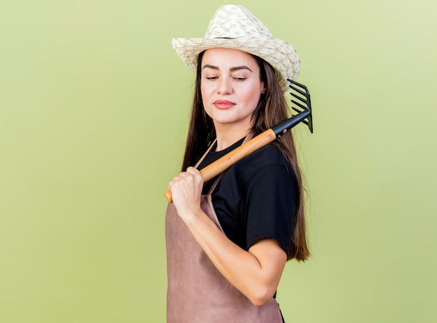 Fiduciosa bella ragazza giardiniere in uniforme che indossa il cappello da giardinaggio mettendo rastrello sulla spalla isolato su sfondo verde oliva