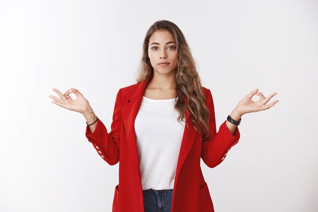 Fiduciosa calma moderna donna d'affari di successo che tiene i sentimenti sotto controllo, mostrando il gesto del mudra della posa del loto in piedi nirvana pacificamente, senza stress, meditando in piedi muro bianco dell'ufficio