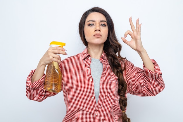 Fiduciosa donna abbastanza caucasica più pulita che tiene in mano un detergente spray e fa un gesto ok segno