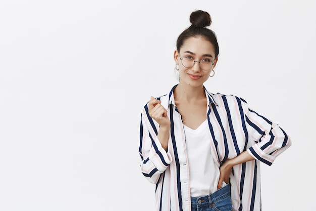 fiduciosa donna affascinante in occhiali e camicia con acconciatura panino, gesticolando con la mano e tenendo il palmo sulla vita, sorridendo, essendo sicura di sé e felice