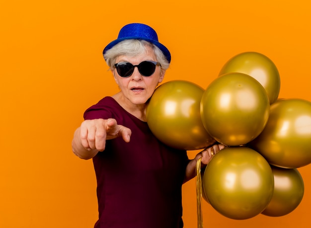 Fiduciosa donna anziana in occhiali da sole che indossano cappello da festa sta con palloncini di elio che punta davanti isolato sulla parete arancione