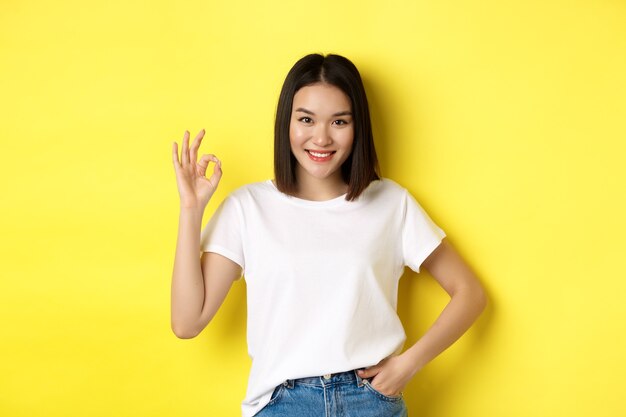 Fiduciosa donna asiatica sorridente e mostrando segno OK, approva e loda la buona offerta, in piedi in maglietta bianca su sfondo giallo.