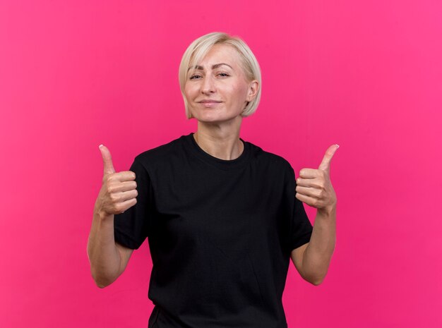 Fiduciosa donna bionda di mezza età guardando la parte anteriore che mostra i pollici in su isolato sulla parete rosa