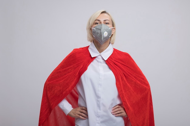 Fiduciosa donna bionda di mezza età supereroe in mantello rosso che indossa una maschera protettiva tenendo le mani sulla vita guardando la parte anteriore isolata sul muro bianco