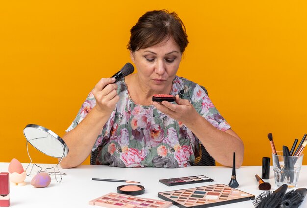 Fiduciosa donna caucasica adulta seduta al tavolo con strumenti per il trucco che tiene il pennello per il trucco e guarda arrossire