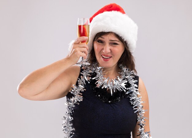 Fiduciosa donna di mezza età che indossa cappello da Babbo Natale e ghirlanda di orpelli intorno al collo alzando un bicchiere di champagne guardando la telecamera isolata su sfondo bianco