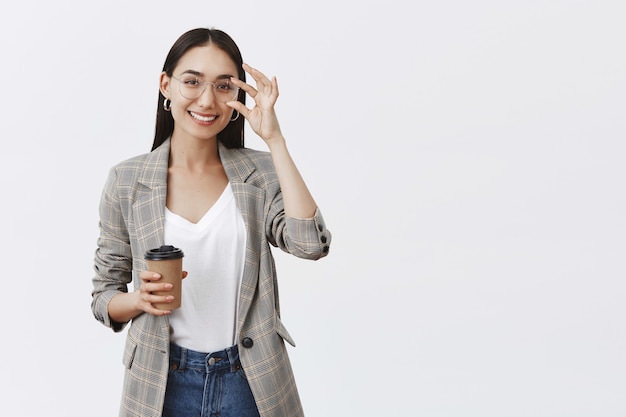 Fiduciosa e gioiosa bella donna in occhiali e giacca elegante, toccando il bordo e sorridendo ampiamente mentre si tiene una tazza di caffè, bevendo bevanda