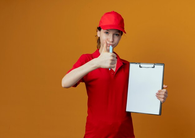 Fiduciosa giovane bella ragazza di consegna in uniforme rossa e cappuccio che tiene penna e appunti e che mostra il pollice in su alla macchina fotografica isolata su priorità bassa arancione con lo spazio della copia