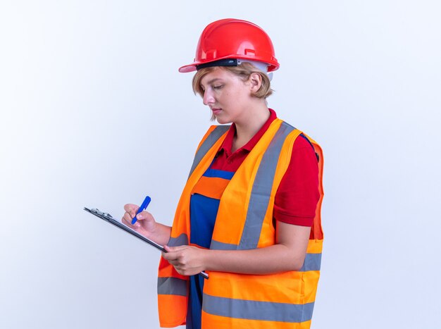 fiduciosa giovane donna costruttore in uniforme che scrive qualcosa negli appunti isolati su muro bianco