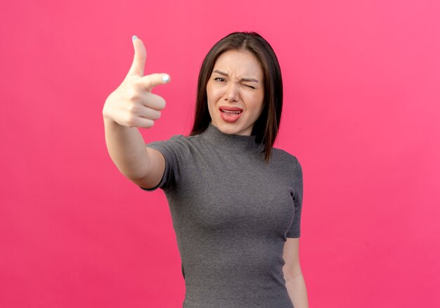 Fiduciosa giovane donna graziosa che sbatte le palpebre e che fa il gesto della pistola alla macchina fotografica isolata su fondo rosa con lo spazio della copia