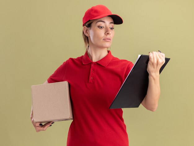 Fiduciosa giovane donna graziosa di consegna in uniforme che tiene cardbox e guardando appunti isolato sulla parete verde oliva