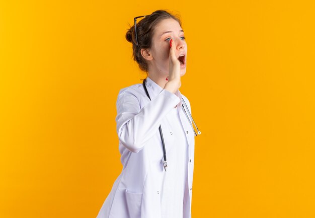 Fiduciosa giovane donna in uniforme da medico con stetoscopio che tiene la mano vicino alla bocca chiamando qualcuno che guarda di lato