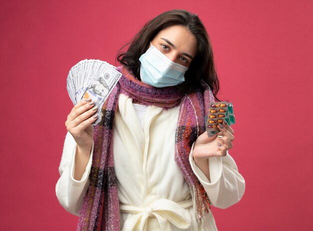 Fiduciosa giovane donna malata che indossa accappatoio e sciarpa con maschera che tiene soldi e confezioni di capsule mediche guardando davanti isolato sul muro rosa