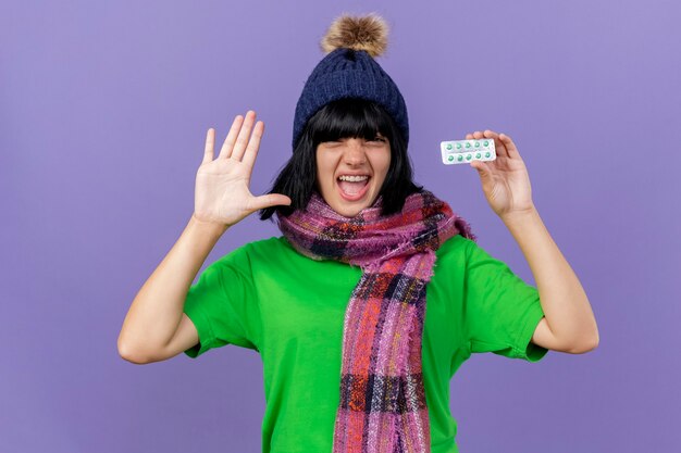 Fiduciosa giovane donna malata che indossa un cappello invernale e sciarpa guardando la confezione anteriore della holding di capsule che mostra la mano vuota isolata sulla parete viola