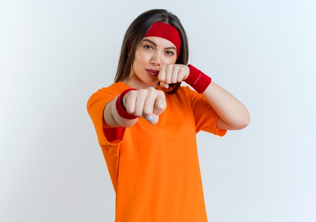 Fiduciosa giovane donna sportiva che indossa la fascia e braccialetti facendo gesto di boxe isolato sulla parete bianca con lo spazio della copia